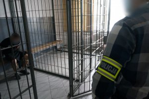 Na zdjeciu zbliżenie na ramie nieumundurowanego policjanta, który ma opaskę odblaskową z napisem policja. W tle widać pomieszczenie dla osób zatrzymanych oraz siedzacego wewnątrz zatrzymanego mężczyznę