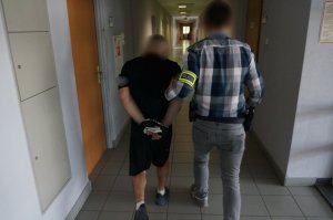 Nieumundurowany policjant prowadzi wewnątrz budynku świętochłowickiej komendy zatrzymnego mężczyznę zakutego w kajdanki na ręce trzymane z tyłu