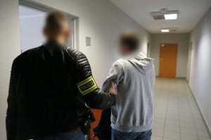 Na zdjeciu niumundurowany policjant z opaska odblaskową na ramieniu z napisem Policja wchodzi do jednego z pomieszczeń służbowych wraz z zatrzymanym mężczyzną