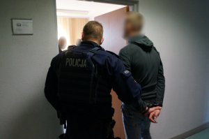 Na zdjeciu umundurowany policjanjacjant oraz zatrzymany mężczyzna zakuty w kajdanki na ręce trzymne z tyłu przed wejściem do jednego z pomieszczeń służbowych