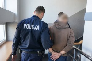 Wnętrze budynku świętochłowickiej komendy. Na zdjęciu umundurowani policjanci prowadzą zatrzymanego mężczyznę zakutego w kajdanki na ręce trzymane z tyłu.