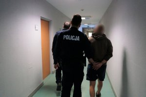 Na zdjęciu umundurowani policjanci prowadzą po korytarzu świętochłowickiej komendy zatrzymanego mężczyznę zakutego w kajdanki na ręce trzymane z tyłu