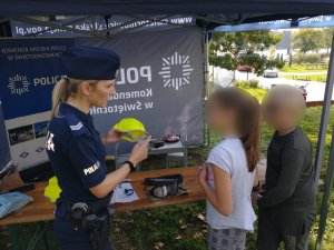 Mundurowa na specjalnie przygotowanym stanowisku rozmawia z dziecmi na temat zagrożeń wynikających w kontakch z obcymi. W rece trzyma żółta czapkę z logiem kampanii Mamo Tato tutaj jestem