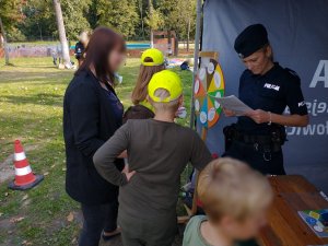 Na zdjęciu widac mundurową która trzyma w dłoniach kartkę z pytania z koła wiedzy. Przy specjalnie przygotowanym stanowiku Policji widać dorosłych i dzieci zaintersowanych zabawą