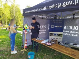 Na zdjęciu widac mundurowego, która trzyma w dłoniach kartkę z pytaniami z koła wiedzy. Przy specjalnie przygotowanym stanowiku Policji widać dwie osoby zaintersowane zabawą