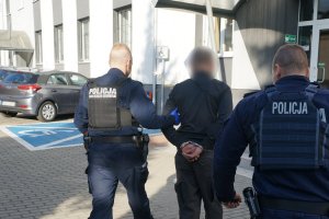 umundurowani policjanci prowadzą zatrzymanego mężczyznę zakutego w kajdanki na ręce trzymane z tyłu do budynku świętichłowickiej komendy