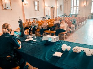 Na pierwszym planie mundurowa siedząca tyłem do obiektywu, prowadząca wykład na temat oszytw. W tle widac siedzacych seniorów, przysłuchujących się prowadzonej prelekcji