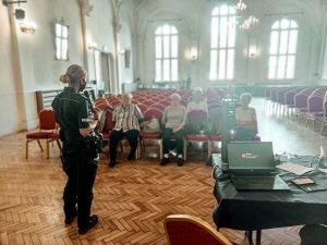 Na zdjęciu na pierwszym planie mundurowa stojąca tyłem do obiektywu, lecz twarzą do słuchaczy. W tle widac siedzacych seniorów, przysłuchujących się prowadzonej prelekcji