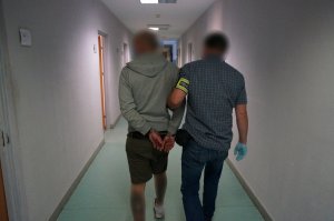 Na zdjęciu nieumunduroway policjant, mający opaskę odblaskową na ramieniu z napisem policja ktory prowadzi po korytarzu świętochłowickiej komendy zatrzymanego mężczyznę zakutego w kajdanki na ręce trzymane z tyłu