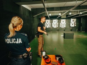 N zdjęciu instruktor objasnia ćwiczenie do wykonania. W dłoni trzyma broń krótką przy pomocy której będzie doskonalił technikę strzelcecką podczas szkolenia z policjantami. Na pierwszym planie widac umundurowaną policjantkę która przysłuchuje się słową instruktora.