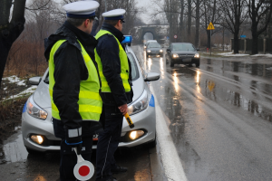 Na zdjeciu dwóch policjntów, za nimi stroi radiowóz. Mundurowi trzymają w ręku tzw. lizaka do wskazania kierującemu miesca zatrzymania oraz urzadzenie do badania trzeźwości. Policjanci obserwuja droge pod kątem kierujących popełnijących wykroczenia. Na zdjęciu widać ze pada deszcz