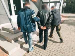 Na zdjeciu nieumundurowani policjanci oznaczeni na ramieniu opaską odblaskową z napisem policja podczas prowadzenia do świętochłowickiej komendy zatrzymanego mężczyznę zakutego w kajdanki na ręce trzymane z tyłu