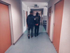 Na zdjęciu korytarz świętochłowickiej komendy. Nieumundurowany policjant prowadzi zatrzymanego mężczyznę zakutego w kajdanki na ręce trzymane z tyłu