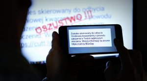 Na zdjęciu zbliżenie na dłonie w ktorych jest telefon oraz treść fałszywego smsa z informacją o kwarantannie w tle widac na ścianie napis o treści oszustwo