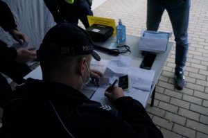 Zbliżenie na policjanta który na stanowisku Stawiennictwa i przyjęcia wpisuje do książki ewidencyjnej rezerwistę który stawił się na ćwiczenia. Policjant trzyma w dłoni dowód osobisty rezerwisty