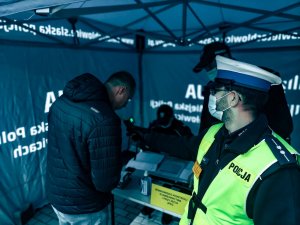 Na zdjeciu wyostrzona postac policjanta w kamizelce odblaskowej ktory przy pomocy urzadzenia do sprawdzania trzeźwości dokonuje badania rezerwisty który stawił się na szkolenie. W tle widac dwóch policjantów którzy wspisują rezerwistę do książki ewidencyjnej.