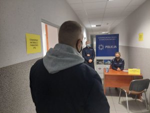 Na zdjęciu widać rezerwiste stojącego plecami do kadru. W tle widać policjantkę siedząca na pnkcie medycznym oraz policjanata ze służby porzadkowo ochronnej