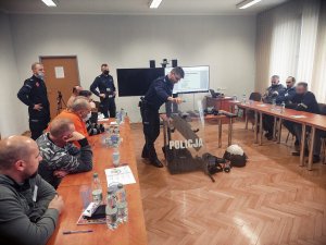 Na zdjęciu policjant przedstawiający rezerwistom wyposażenie i sprzęt policyjny, który rozlożony został na stoliku znajdującym się pośrodku sali