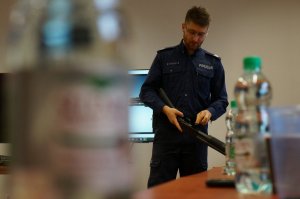 Na zdjeciu zblienie na policjanta który trzyma w ręce broń długa typu mosberg. Policjant objaśnia w jaki sposób załoadować amunicję