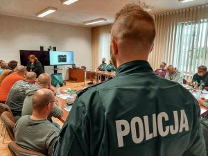 Na zdjęciu zbliżenie na napis policja znajdujący się na bluzie ćwiczebnej policjanta. W tle widać siedzacych za stołami rezerwistów oraz prowadzącą wykład z pierwszej pomocy.