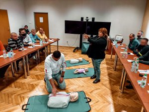 Na zdjeciu sala wykładowa w której za stolikami siedzą rezerwiści. Na środku sali widać ochotnika klęczącego przy fantomie oraz prowadzacą wykład, która pokzuje jak właściwie układać ręce do przeprowadzenia masażu serca