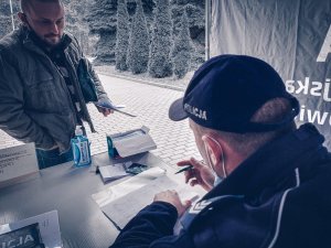 Scena przedstawia rezerwiste stojącego przy stoliku punktu kontroli i stawiennictwa i przyjęcia po zakończonych ćwiczeniach. Policjant odnotowuje w książce godzinnę wyjścia rezerwisty po zakończonych ćwiczeniach