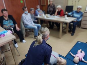 Na zdjęciu policyjna profilaktyk omawia seniorom którzy siedzą przy stałach zasady udzielania pierwszej pomocy. Policjantka klęczy na ziemi, przed nią leży fantom odzwierciedlający postac dorosłej osoby.