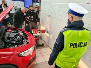 Na zdjęciu prawej stronie umndurowany policjant w odblaskowej kamizelce. Przed nich stoi zaparkowany pojazd, przy ktorym pracownik stacji diagnostycznej sprawdza światła. Na zdjęciu widać także mężcyznę z kamerą który rejestruje całe zdarzenie.