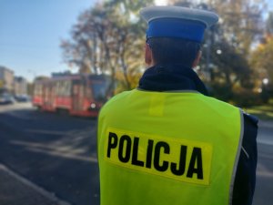 Na zdjęciu widać policjana w żółtej kamizelce odblaskowej policja. Zbliżenie na napis w tle widac przejeźdzający tramwaj