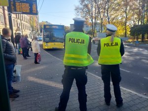 Na zdjęciu widać policjantkę i policjanta z drogówki oraz pasażerów ocekujących na przyjazd autobudu. Mundurowi oczekują na autobus w celu przeprowadzenia kontroli w związku z obowiązkiem zakrywania ust i nosa.