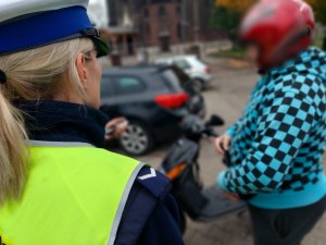 Na zdjeciu zbliżenie na pagon sierżnta na ramieniu policjanci z dorgówki, która prowadzi kontrolę drogową kierującego motorowerem. W tle widać motorower i postac kontrolowanego mężczyzny