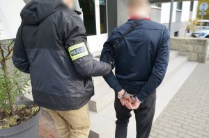 Na zdjęciu nieumundurowany policjant z założoną na ramieniu opaską odblaskową z napisem Policja prowadzi zatrzymanego mężczuznę zakutego w kajdanki na ręce trzymane z tyłu do budynku świętochłowickiej komendy