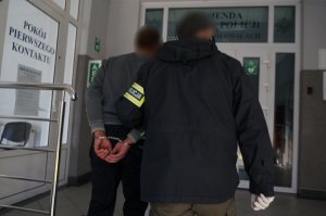 Na zdjęciu nieumundurowany policjant z założoną na ramieniu opaską odblaskową z napisem Policja prowadzi zatrzymanego mężczuznę zakutego w kajdanki na ręce trzymane z tyłu