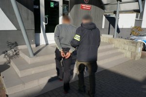 Na zdjęciu nieumundurowany policjant z założoną na ramieniu opaską odblaskową z napisem Policja prowadzi zatrzymanego mężczuznę zakutego w kajdanki na ręce trzymane z tyłu do budynku świętochłowickiej komendy