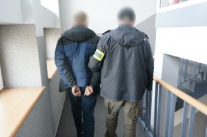 Na zdjęciu nieumundurowany policjant z opaską odblaskową z napisem policja prowadzi zatrzymanego mężczyznę zakutego w kajdanki na ręce trzymane z tyłu schodami na wyższa kondygnację budynku