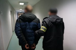 Na zdjęciu nieumundurowany policjant z opaską odblaskową z napisem policja prowadzi korytarzem zatrzymanego mężczyznę zakutego w kajdanki na ręce trzymane z tyłu