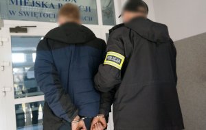 Na zdjęciu nieumundurowany policjant z opaską odblaskową z napisem policja prowadzi zatrzymanego mężczyznę zakutego w kajdanki na ręce trzymane z tyłu do budynku komendy