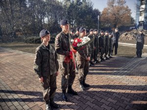 Na zdjeciu stojących w dwu szeregu kadetów. Przed nimi troje uczniow ustawionych w poczet honorowy z wiązanką kwiatów w rękach.