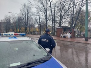 Na zdjeciu policjant w trakcie obserwacji sytuacji na cmentarzu podczas Święta Zmarłych, na pierwszym planie widać napis policja na belce oświetleniowej radiowozu