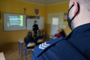 Zbliżenie na pagon starszego sierżanta, znajdujący się na ramieniu policjanta. w tle widać postac policjantki podczas rozmawiania z młodzieżą na temat odopowiedzialności prawnej osób nieletnich