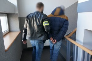 Na zdjęciu wnętrze budynku Komendy Miejskiej Policji w Świętochłowicach. Nieumundurowany policjant z opaska odblaskową na ramieniu z napisem Policja prowadzi zatrzymanego mężczyznę zakutego w kajdanki na ręce trzymane z tyłu