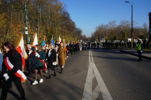 Na zdjęciu widac kolumnę pieszych składająca się z mieszkańców miasta, władz miejskich, pocztów sztandarowych, reprezentantów służb mundurowych oraz licznych delegacji,  którzy maszerują jedną ze świętochłowickich ulic. Na zdjęciu widać mundurową która czuwa nad bezpieczeństwem uczestników