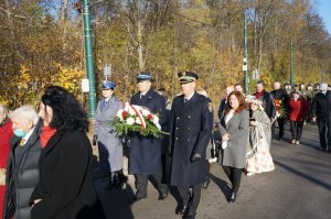 Na zdjęciu widac kolumnę pieszych składająca się z mieszkańców miasta, władz miejskich, pocztów sztandarowych, reprezentantów służb mundurowych oraz licznych delegacji,  którzy maszerują jedną ze świętochłowickich ulic. Zbliżenie na Pierwszego Zastepcę Komendanta Miejsckiego Policji w Świętochłowicach oraz Komendanta Strazy Miejskiej i Komendanta Państwowej Straży Pożarnej który niesie wieniec