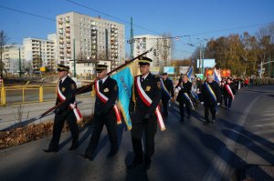 Na zdjęciu widac kolumnę pieszych składająca się z mieszkańców miasta, władz miejskich, pocztów sztandarowych, reprezentantów służb mundurowych oraz licznych delegacji,  którzy maszerują jedną ze świętochłowickich ulic. Na pierwszym planie poczet sztandarowy składający się z trzech strażników miejskich