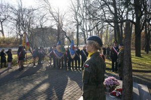 Na zdjęciu wiadac dwie kadetki stojące przy pomniku Powstańców Śląskich, w tle widać ustawiające się obok siebie osoby bedące w pocztach sztandarowych różnych instytuacji