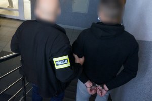 Na zdjęciu nieumundurowany policjant prowadzi zatrzymanego mężczyznę zakutego w kajdanki na ręce trzymane z tyłu schodami w dół do wyjścia z komendy