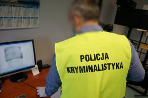 Zdjęcie zrobione za pleców policjanta ubranego w kamizelkę odblaskową z napise Policja Kryminalistyka podczas pobierania od zatrzymanego mężczyzny odcisków palca. W tle widać monitor na którym widac zarys linii papilarnych