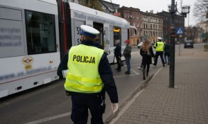 Na zdjęciu widać stojący na przystanku tramwaj. Obok widać ludzi oraz mundurowych zmierzających do tramwaju. Umundurowani policjncji wraz z pracownicami sanepidu przeprowadzenią kontrolę stosowania się przed podróżnych do obostrzeń