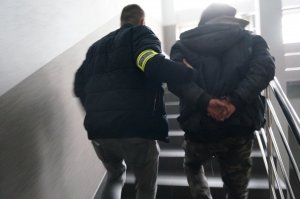 Na zdjęciu nieumundurowany policjant, który ma na ramieniu założoną opaskę odblaskową z napisem Policja prowadzi schodami na wyższe piętro budynku świętochłowickiej komendy zatrzymanego mężczyznę zakutego w kajdanki na ręce trzymane z tyłu