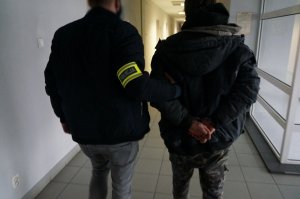 Na zdjęciu nieumundurowany policjant, który ma na ramieniu założoną opaskę odblaskową z napisem Policja prowadzi korytarzem w budynku świętochłowickiej komendy zatrzymanego mężczyznę zakutego w kajdanki na ręce trzymane z tyłu